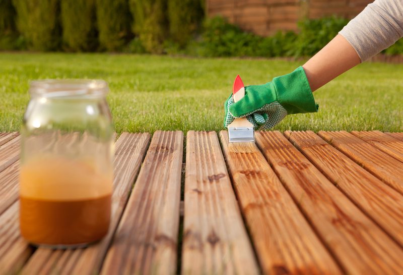 Person in a green glove uses a small roller to stain wooden deck boards; a jar of brown stain sits on the deck with a green lawn in the background.