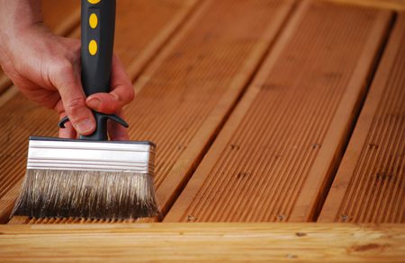 Close-up of a hand holding a wide paintbrush with a black handle applying stain to parallel wooden deck boards.