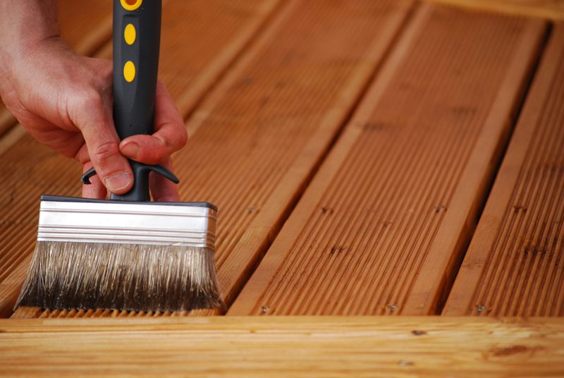Close-up of a hand holding a wide paintbrush with a black handle applying stain to parallel wooden deck boards.
