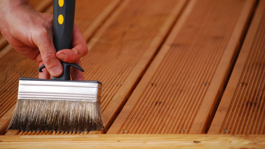 Close-up of a hand holding a wide paintbrush with a black handle applying stain to parallel wooden deck boards.