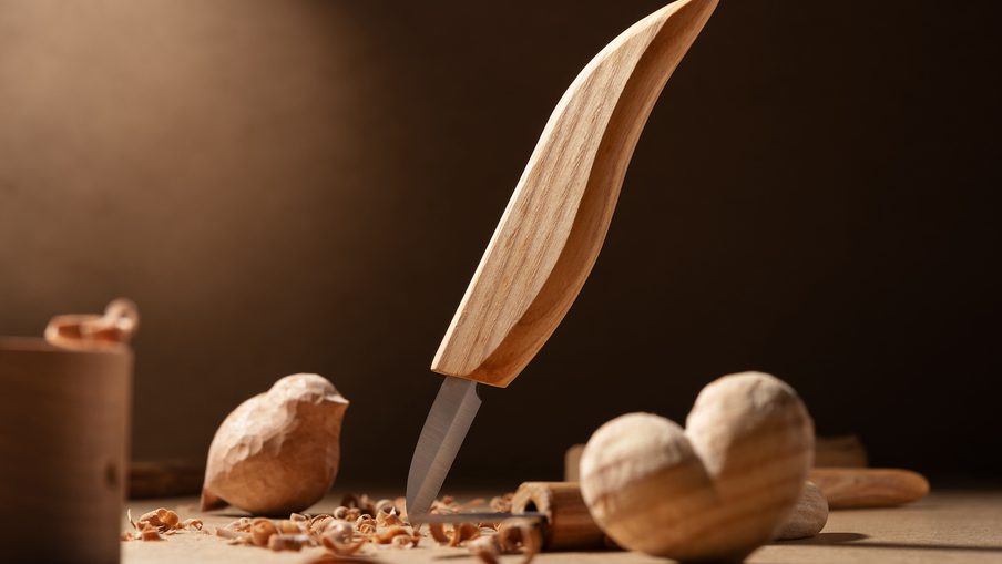 Warm woodworking scene with a curved wooden-handled carving knife standing upright in a small wooden block, shavings and turned pieces on the workbench