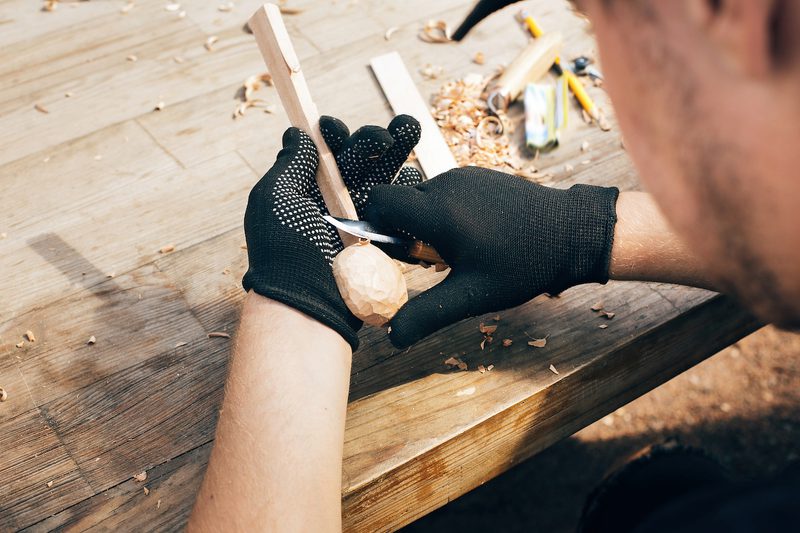 A person wearing black gloves carves a wooden bead on a workbench, using a knife; wood shavings and carving tools are scattered nearby.