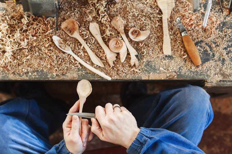 Overhead view of a person carving a wooden spoon at a bench, with wood shavings and carving tools scattered, hands guiding a blade.
