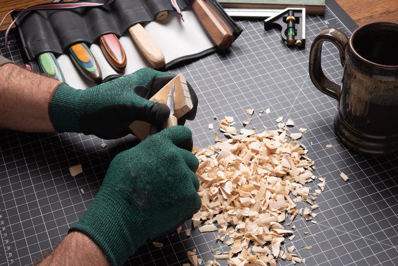 Hands in green gloves carve a wooden block on a grid cutting mat, with wood shavings and tools nearby.