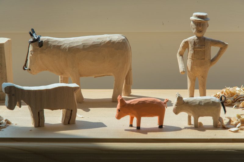 Wooden carved figures on a light wooden surface with shavings: a person wearing a hat stands with hands on hips beside animals, including a horse and pig.