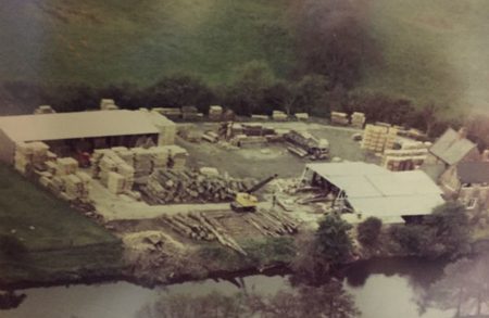 Aerial view of a lumber yard by a river: stacked timber, pallets, a white-roofed work area, sheds, equipment, and trees in the background.