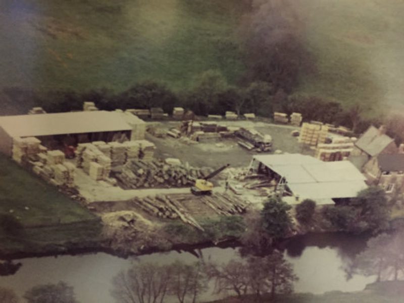 Aerial view of a lumber yard by a river: stacked timber, pallets, a white-roofed work area, sheds, equipment, and trees in the background.
