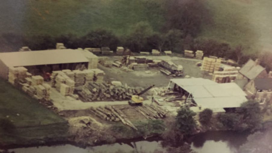 Aerial view of a lumber yard by a river: stacked timber, pallets, a white-roofed work area, sheds, equipment, and trees in the background.