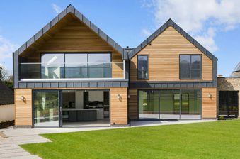 Modern two-story house with wood siding, large glass sliding doors and windows, a glass balcony, dark metal trim, and a green lawn under a blue sky.