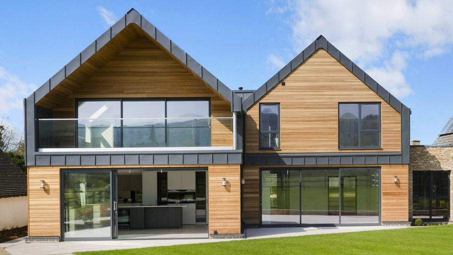 Modern two-story house with wood siding, large glass sliding doors and windows, a glass balcony, dark metal trim, and a green lawn under a blue sky.
