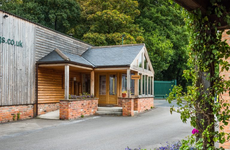 Small brick-and-wood showroom with glass walls and a wooden door, beside a larger wooden building. Ivy-covered brick column, fence, and trees behind.