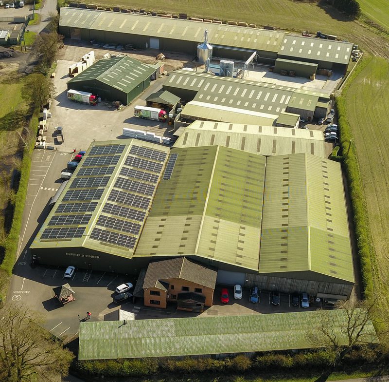 An aerial view of a large industrial complex with green metal roofs, some solar panels, loading bays, parked trucks and cars, bordered by grass fields.