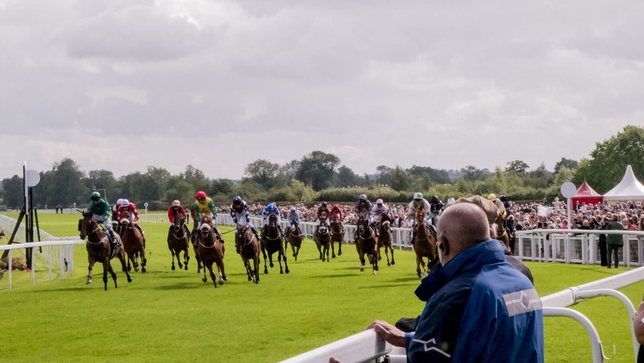 Jockeys race horses on a green turf track, crowd behind white rails, under a cloudy sky; a spectator in a blue jacket watches from the foreground.