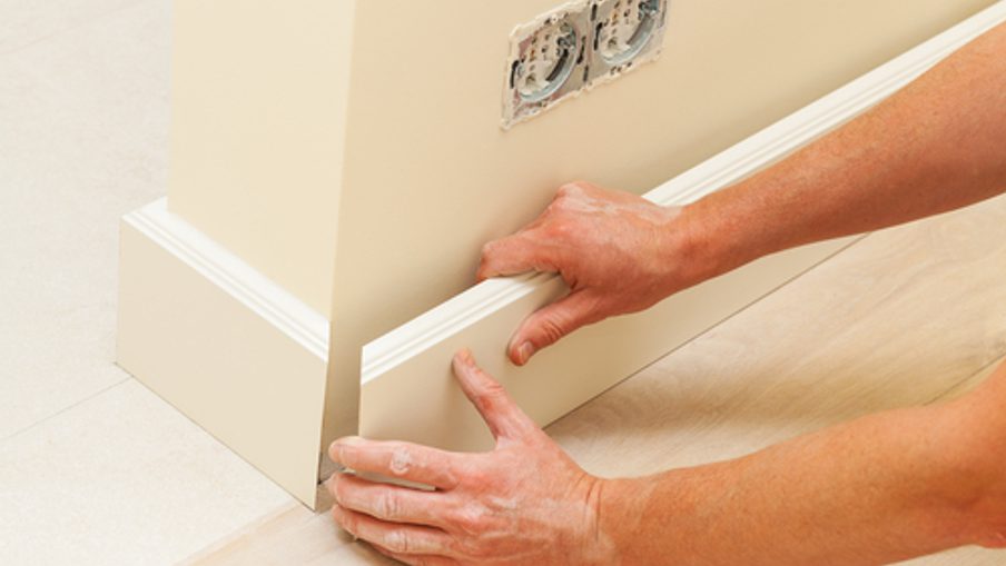 Close-up of hands installing a white baseboard along a wall, with an electrical outlet visible and light-colored flooring.