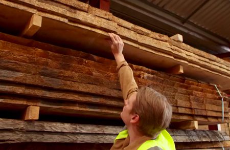 Person in a neon safety vest reaches up to touch the underside of a top plank in a stacked lumber pile inside a warehouse.