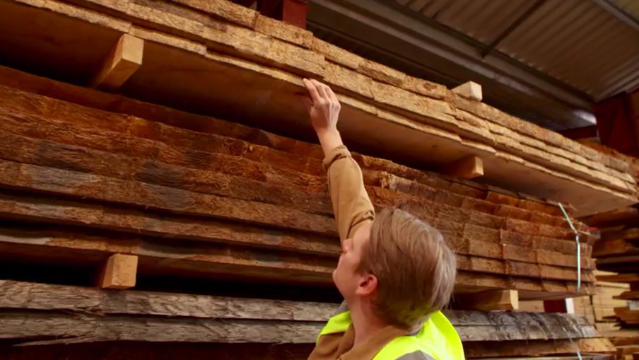 Person in a neon safety vest reaches up to touch the underside of a top plank in a stacked lumber pile inside a warehouse.