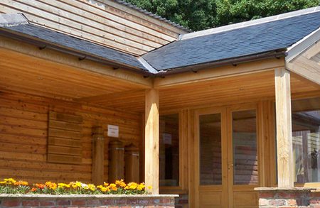 Wooden timber building with a covered porch and glass conservatory, brick base, a flower planter with orange blooms, and a circular logo in the top-right.
