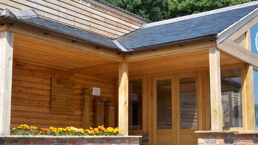 Wooden timber building with a covered porch and glass conservatory, brick base, a flower planter with orange blooms, and a circular logo in the top-right.