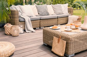 Cozy outdoor lounge on a deck with a woven rattan sofa, neutral cushions, a matching coffee table with cups and pastries, and wicker baskets nearby.