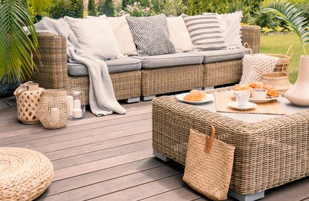 Cozy outdoor lounge on a deck with a woven rattan sofa, neutral cushions, a matching coffee table with cups and pastries, and wicker baskets nearby.