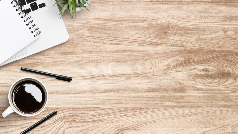 Desk setup with a laptop, spiral notebook, small plant, two black pens, and a cup of black coffee on a light wood surface.
