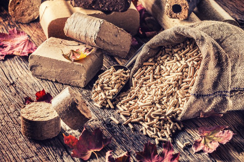 Burlap sack spilling wood pellets onto a weathered wooden surface, with autumn leaves and chunky briquettes nearby.