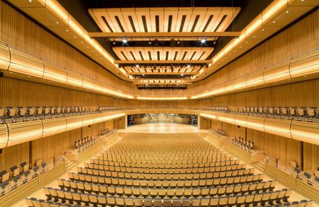 Empty modern concert hall with warm wooden walls, tiered balconies on both sides, a distant stage, and orderly rows of beige seats filling the auditorium.