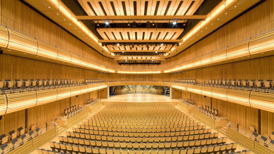 Empty modern concert hall with warm wooden walls, tiered balconies on both sides, a distant stage, and orderly rows of beige seats filling the auditorium.