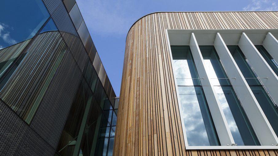 Low-angle view of a modern building with curved vertical wooden cladding and tall glass windows with white frames against a blue sky.