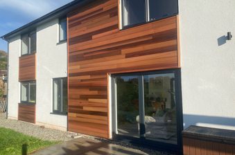 Modern two-story home with white plaster and horizontal wooden cladding in varying brown tones. Glass sliding doors, gravel path, and blue sky.