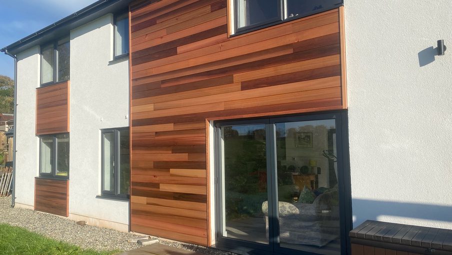 Modern two-story home with white plaster and horizontal wooden cladding in varying brown tones. Glass sliding doors, gravel path, and blue sky.