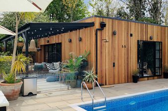 Wooden poolside cabin with glass walls, string lights, and outdoor seating among potted plants; blue-tiled pool in the foreground.