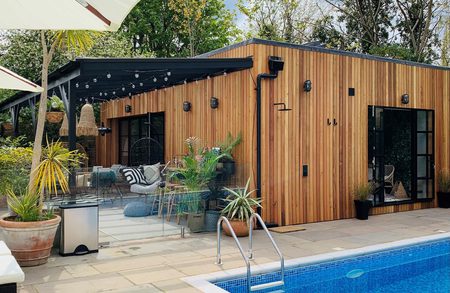 Wooden poolside cabin with glass walls, string lights, and outdoor seating among potted plants; blue-tiled pool in the foreground.