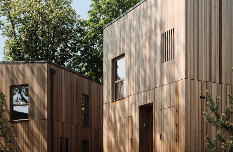 Two modern wooden houses with vertical timber siding and large windows, connected by a wooden deck, set among trees under a clear blue sky.