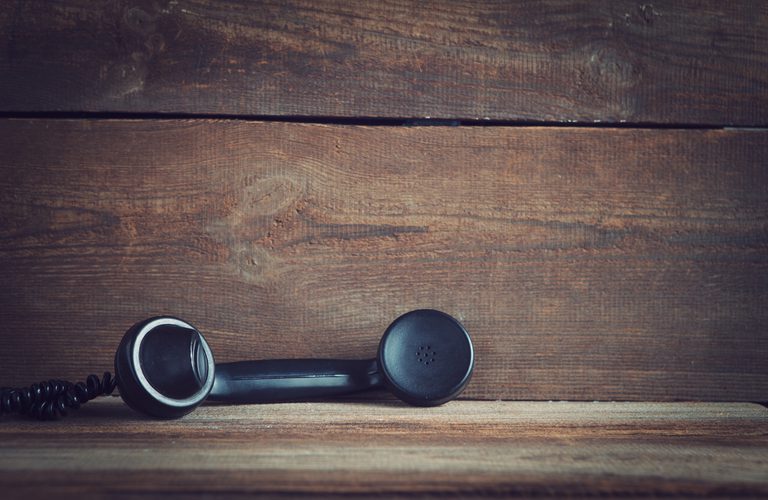 An old-fashioned black corded telephone handset rests on a weathered wooden surface with a wood-paneled background; a coiled cord trails to the left.