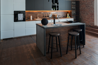 Industrial loft kitchen with dark wood floors, grey cabinetry, brick wall, large grid window, and a central island with three stools.