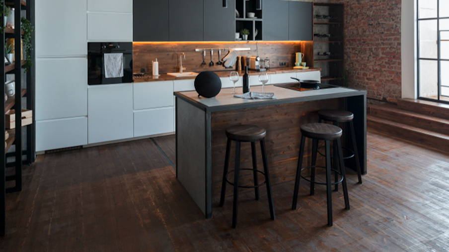 Industrial loft kitchen with dark wood floors, grey cabinetry, brick wall, large grid window, and a central island with three stools.