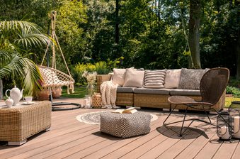 Outdoor wicker patio seating area on a wooden deck, featuring a sofa with cushions, a hanging chair, patterned pouf, plants, lanterns, and a circular rug.