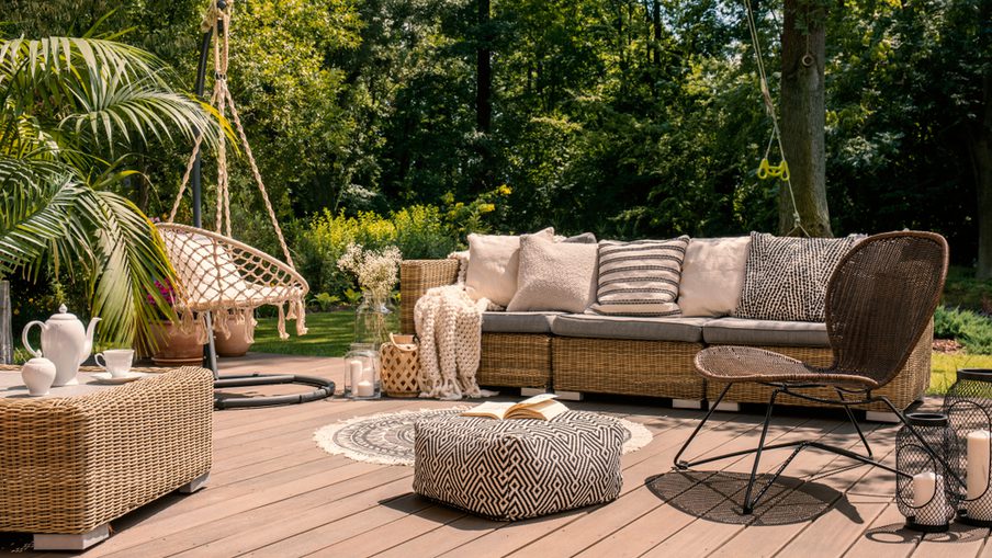 Outdoor wicker patio seating area on a wooden deck, featuring a sofa with cushions, a hanging chair, patterned pouf, plants, lanterns, and a circular rug.