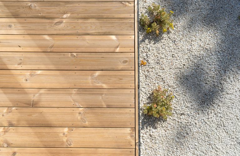 Left: wooden deck boards; right: light gravel with small shrubs; a shadow of a person falls on the gravel.