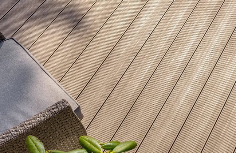 Top-down view of a brown wicker outdoor chair with a gray cushion on a light wooden deck, with green leaves in the bottom corner.