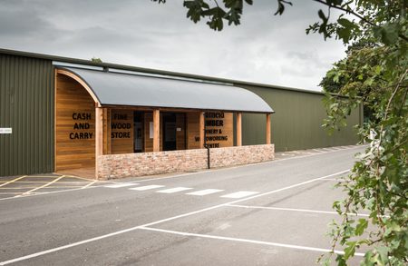 Timber and wood store with a curved metal canopy, brick low wall, and CASH AND CARRY sign, beside a green warehouse and a street with crosswalk markings.