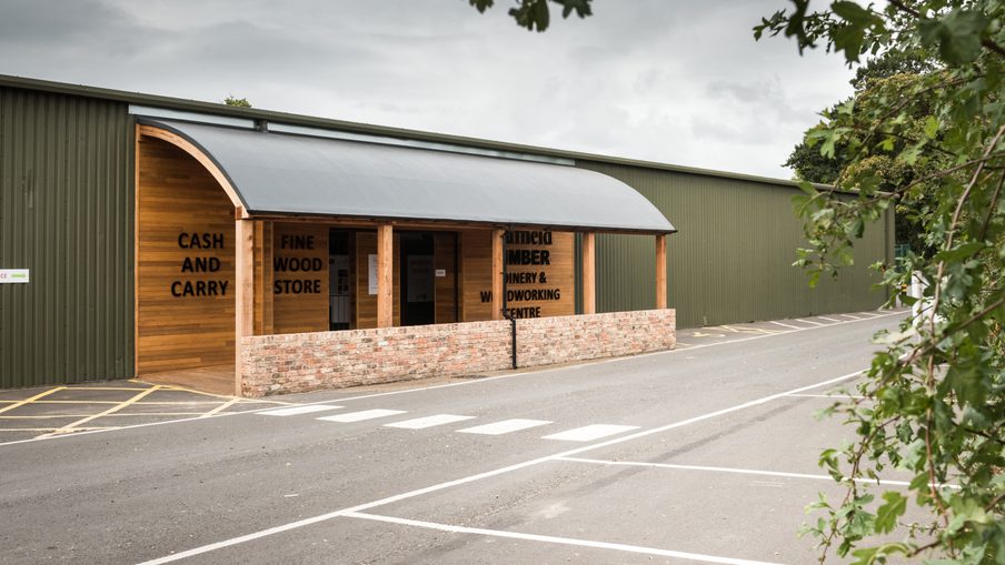 Timber and wood store with a curved metal canopy, brick low wall, and CASH AND CARRY sign, beside a green warehouse and a street with crosswalk markings.