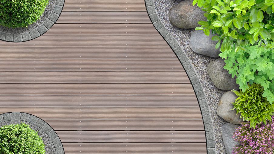 Top-down view of a curved wooden deck path edged with gray stones, with rocks and lush green shrubs on the right and round planters on the left.