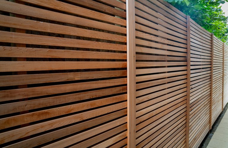 Long privacy fence of evenly spaced horizontal wooden slats with vertical posts along a walkway; greenery visible at the top.