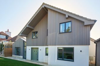 Modern two-story house with gray vertical siding over a white base, large windows, and a steep sloped roof; stone patio and green lawn in front.