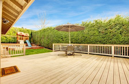 Backyard wooden deck with a round table and umbrella, bordered by tall green hedges; a playset with a slide sits to the left under a bright blue sky.