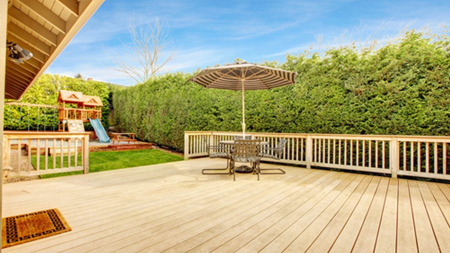 Backyard wooden deck with a round table and umbrella, bordered by tall green hedges; a playset with a slide sits to the left under a bright blue sky.
