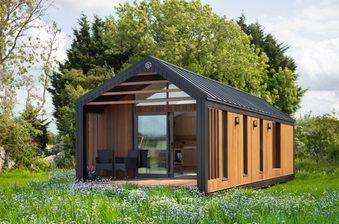 Modern wooden cabin with a black metal frame and gabled roof, glass sliding door, open porch with chairs, in a green meadow with wildflowers and trees.