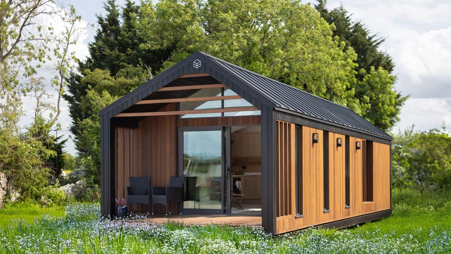 Modern wooden cabin with a black metal frame and gabled roof, glass sliding door, open porch with chairs, in a green meadow with wildflowers and trees.